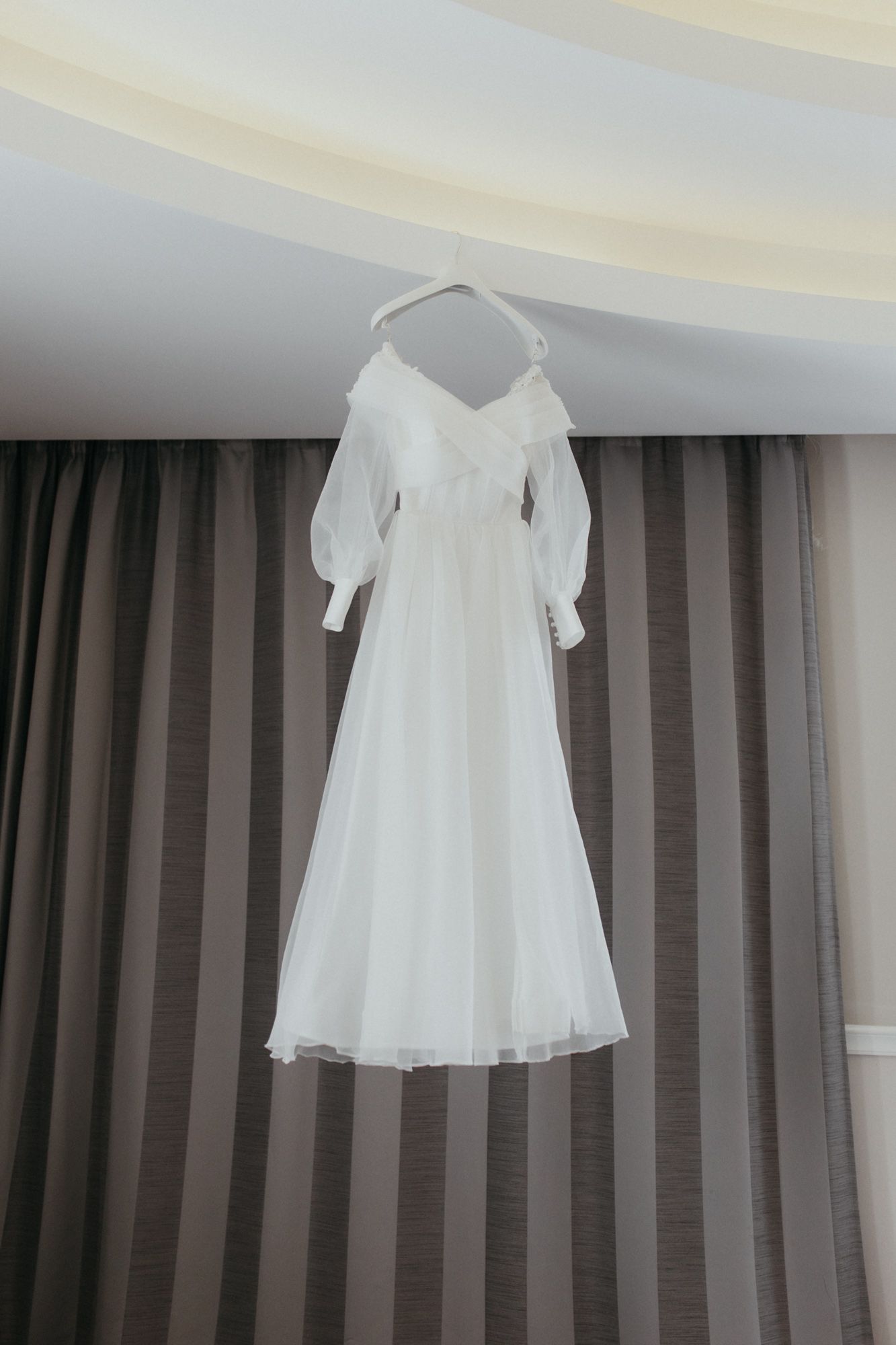 Elegant white wedding dress with long sleeves hanging against gray striped curtains.