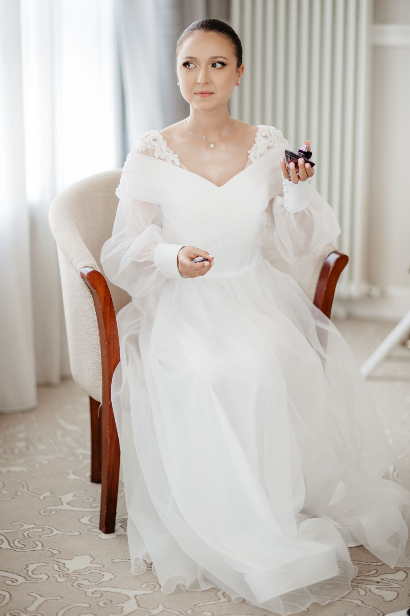 Bride in elegant white wedding dress with puffy sleeves sitting in upholstered chair against light curtains.