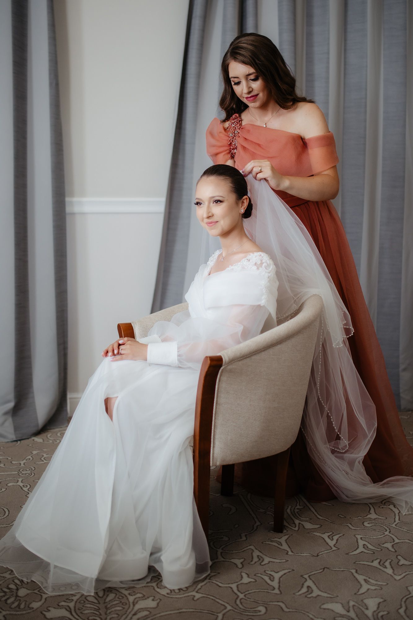 Wedding preparation scene with bride in white dress seated while attendant in coral dress helps with veil.