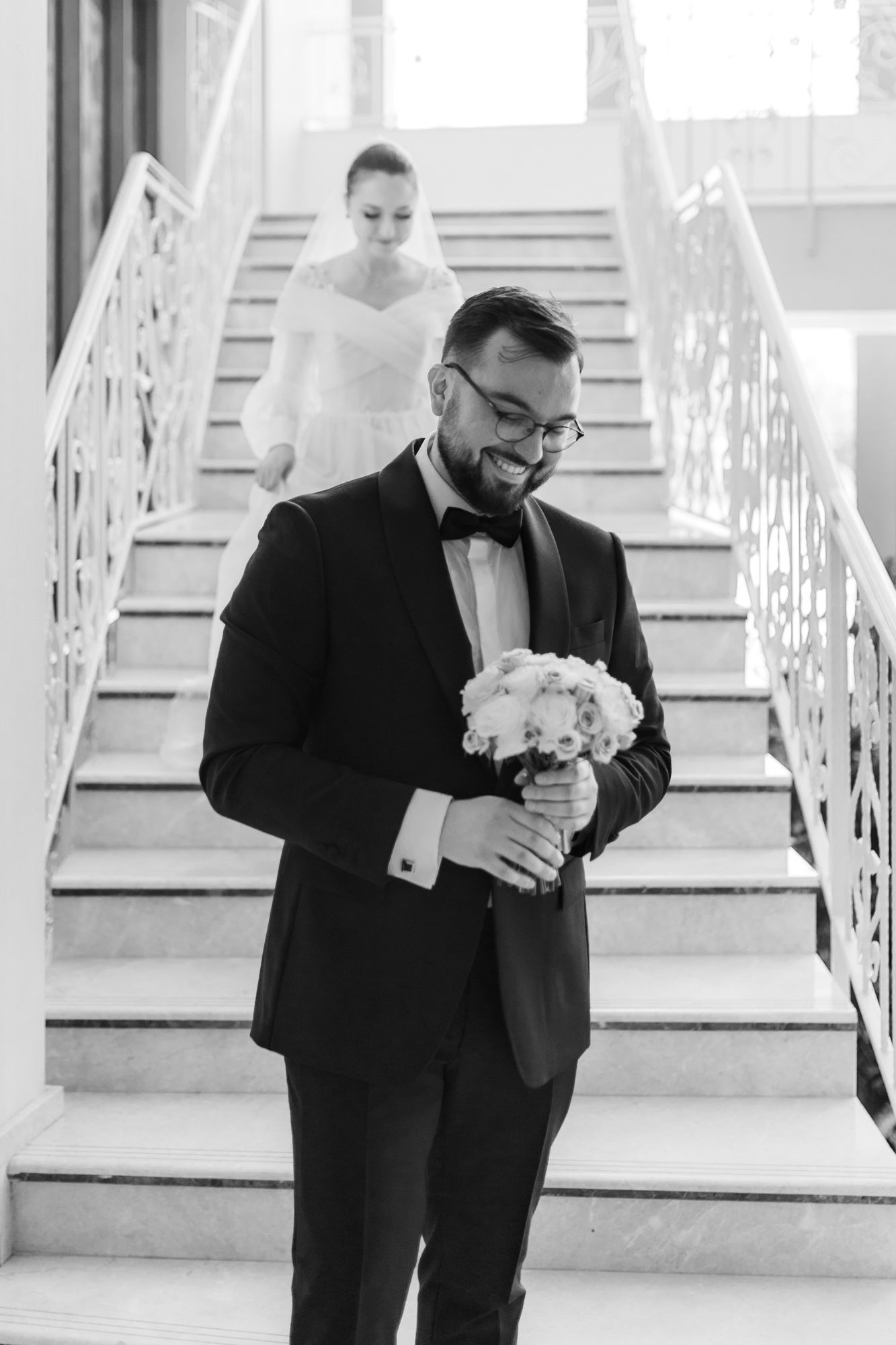 Black and white photo of groom holding bouquet on stairs with bride in background.