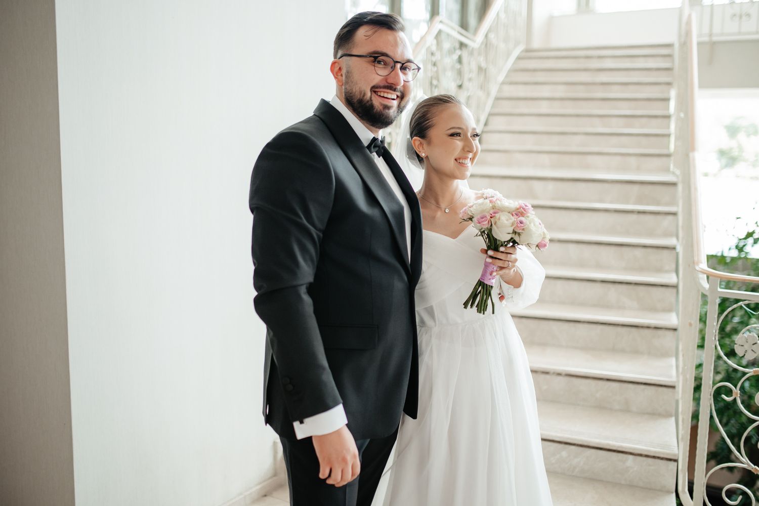 Newlywed couple posing together on elegant white staircase with ornate railings.