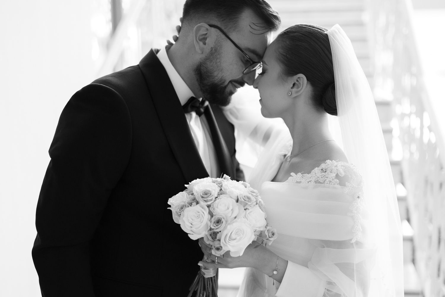 A romantic black and white wedding photo shows a tender moment as the couple touches foreheads with white roses in hand.