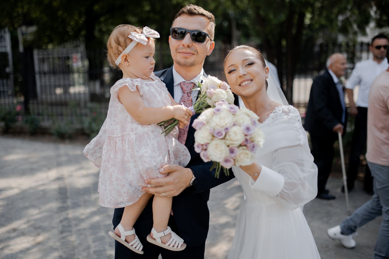 A joyful wedding party poses outdoors with a small child and pink floral bouquets on a sunny day.