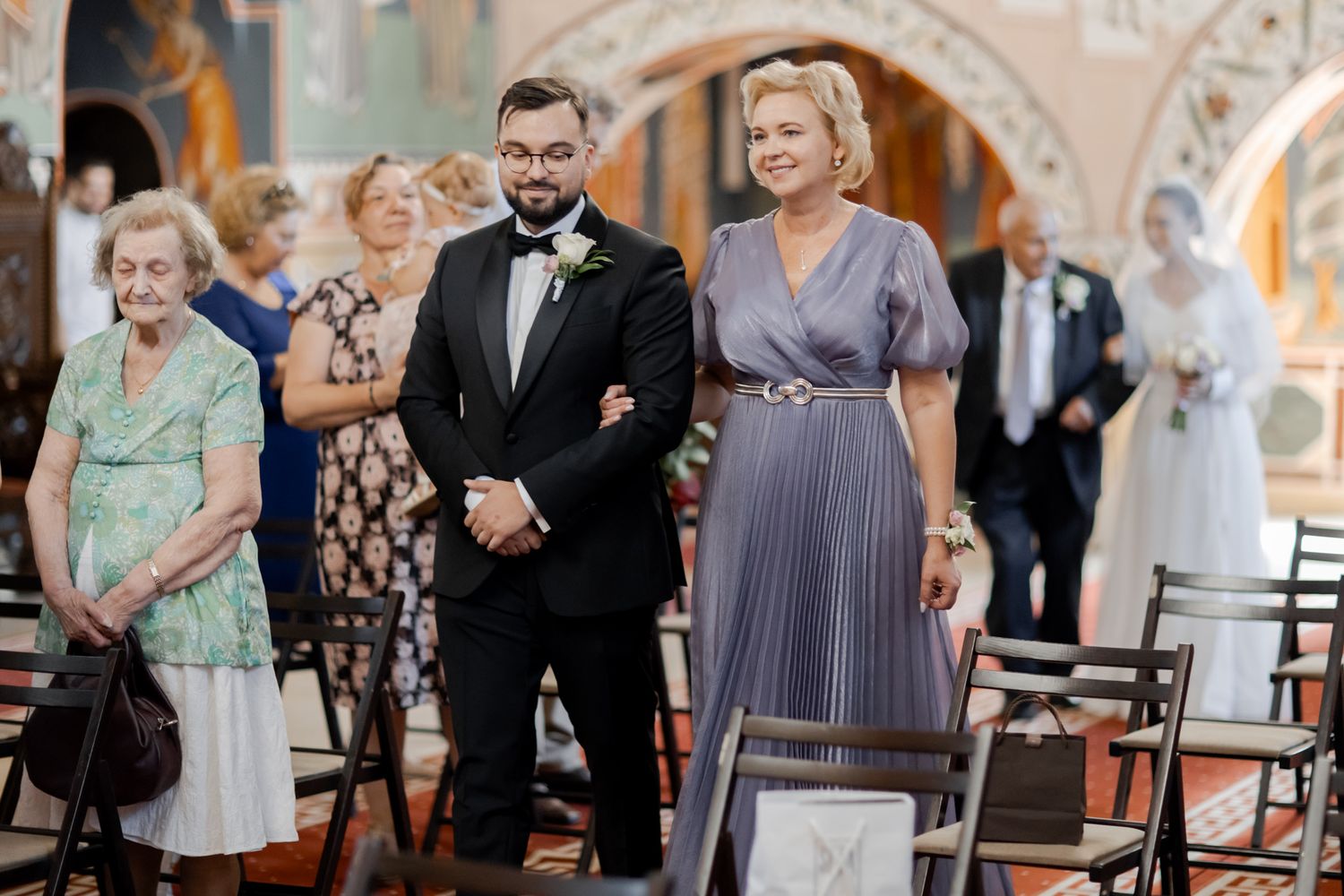 Wedding ceremony guests stand in an ornate church interior with arched architecture and decorative details.