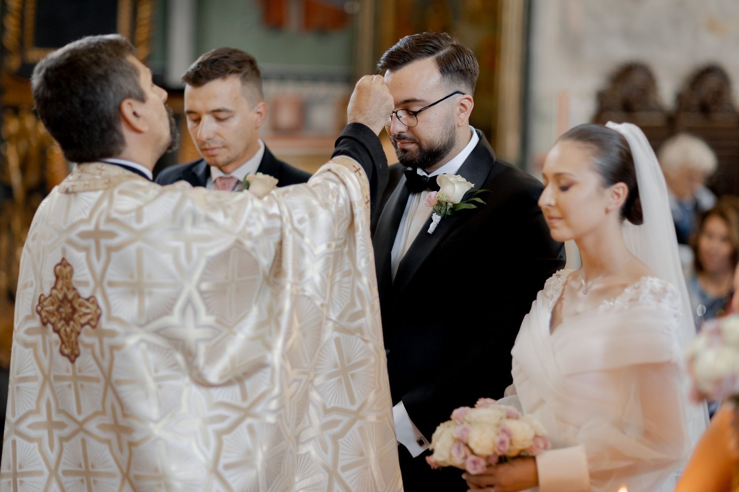 Orthodox wedding ceremony taking place in an ornate church with priest in white robes blessing the couple.