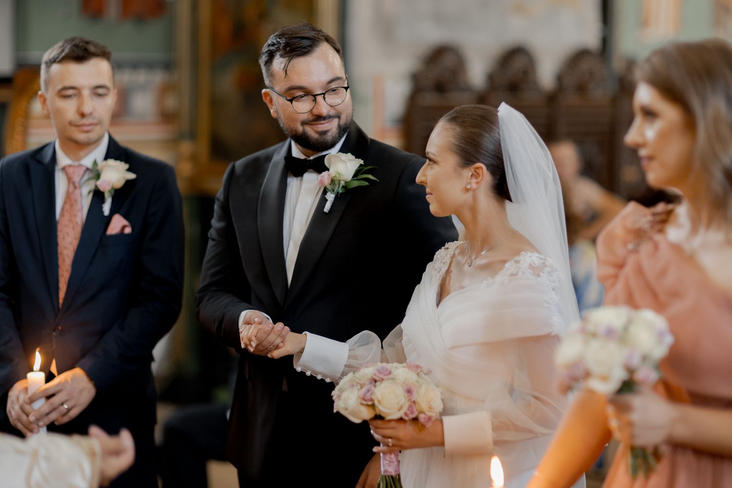 Wedding party gathered during Orthodox ceremony with lit candles and floral arrangements.