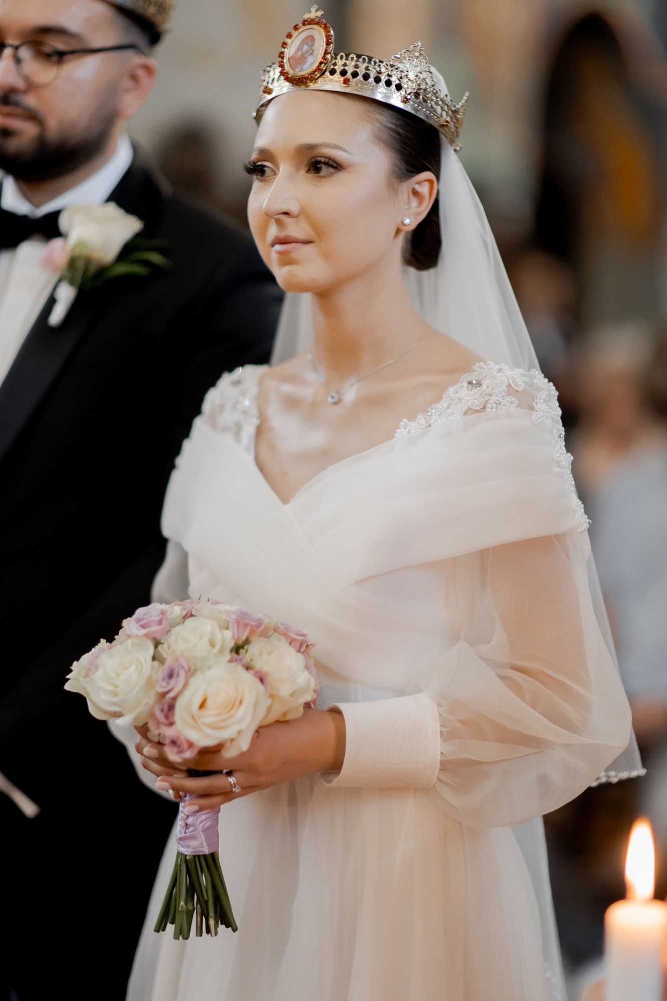 Bride in elegant white dress and crown holds bouquet of cream roses during orthodox ceremony.