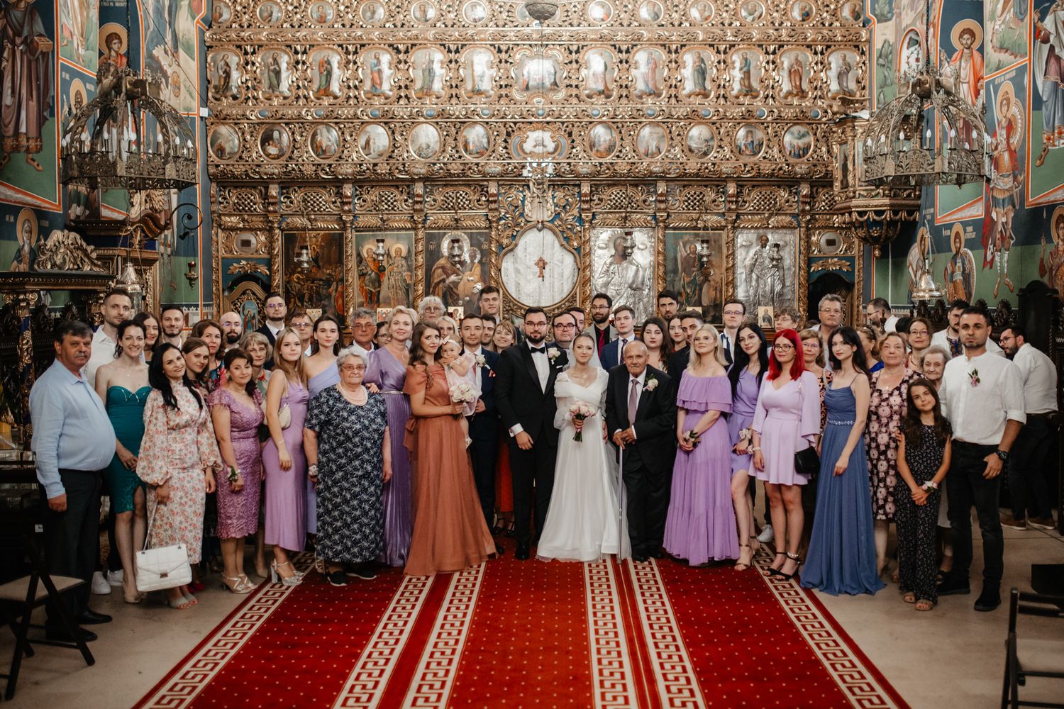 Large wedding party photo in orthodox church with ornate iconostasis and guests in formal attire.