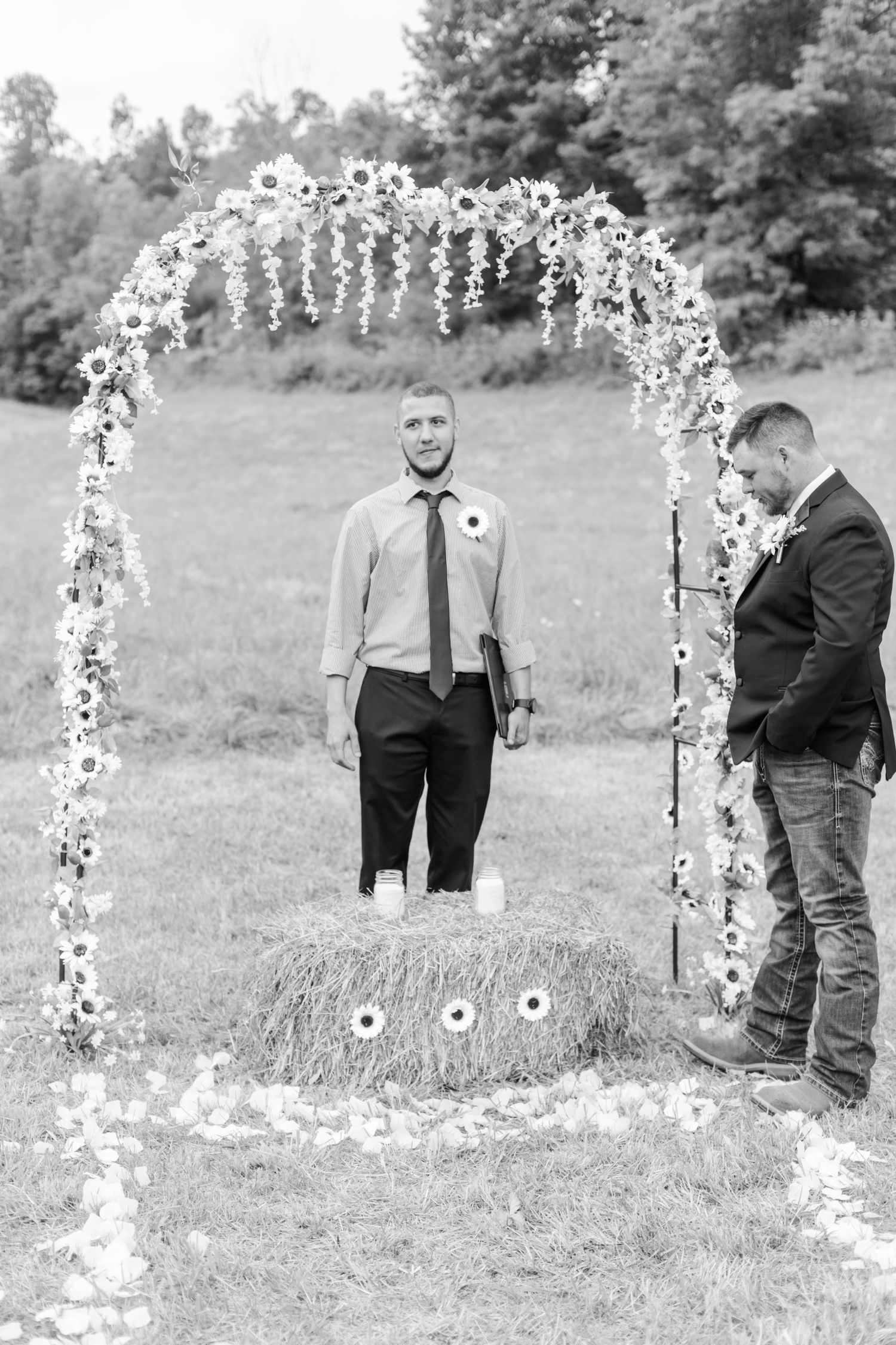 Black and white photo of wedding party members standing near a floral-decorated arch outdoors.
