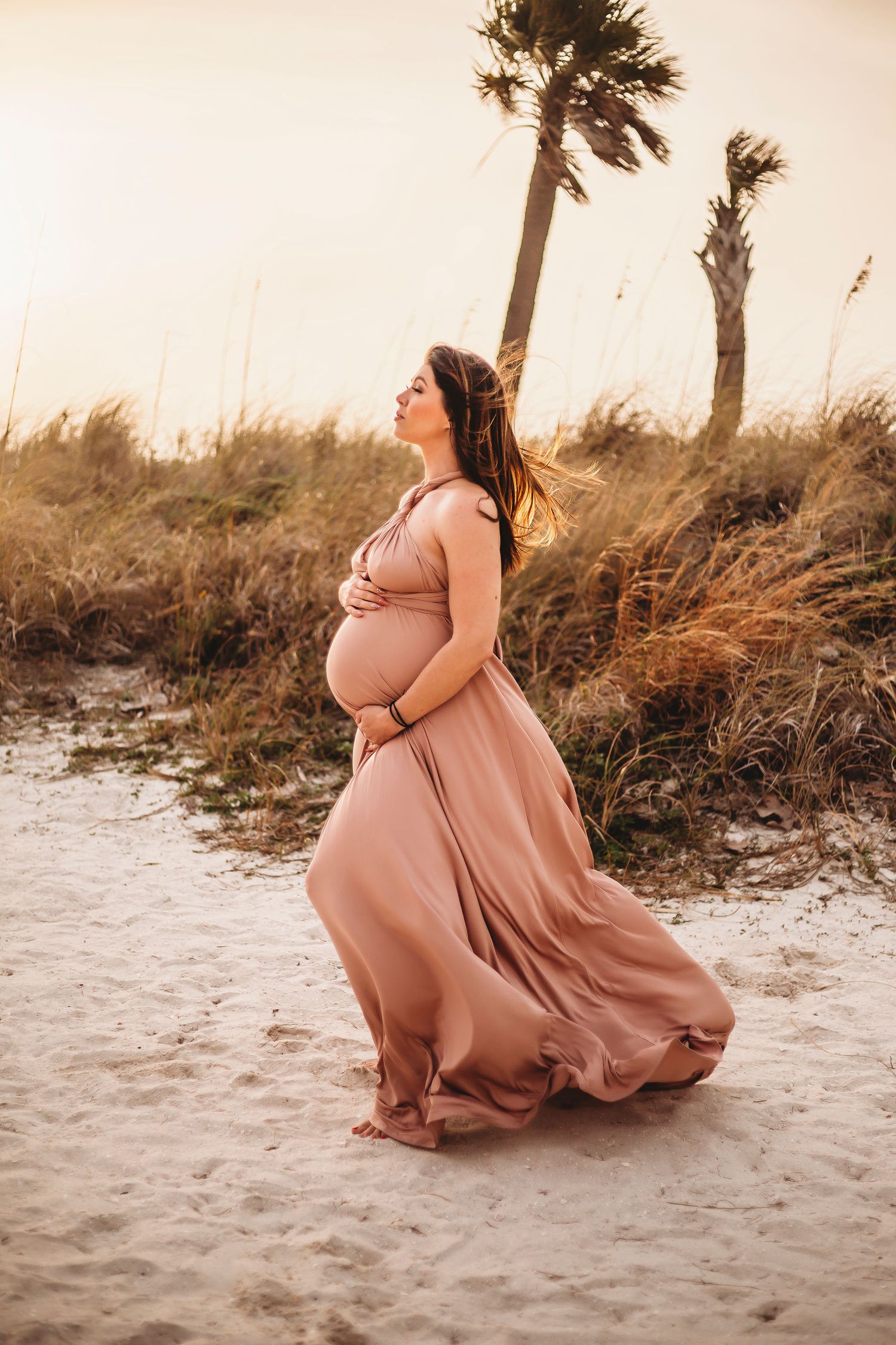 Expectant mother in flowing pink dress poses on beach at sunset with palm trees.