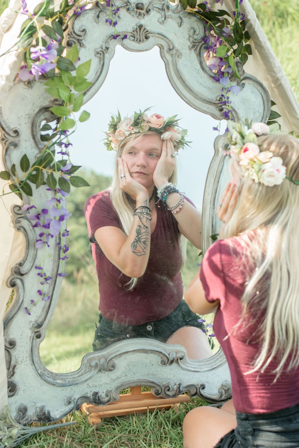 An ornate mirror decorated with purple flowers and greenery sits in a sunny meadow.