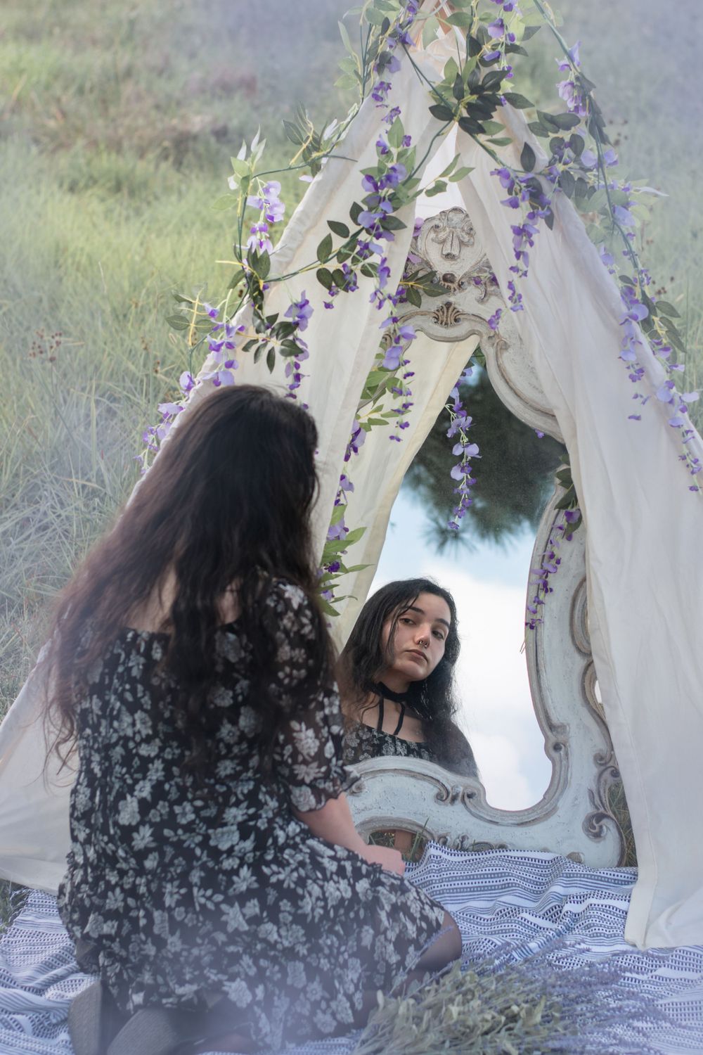 A dreamy outdoor scene with an ornate mirror decorated with purple flowers and white fabric.