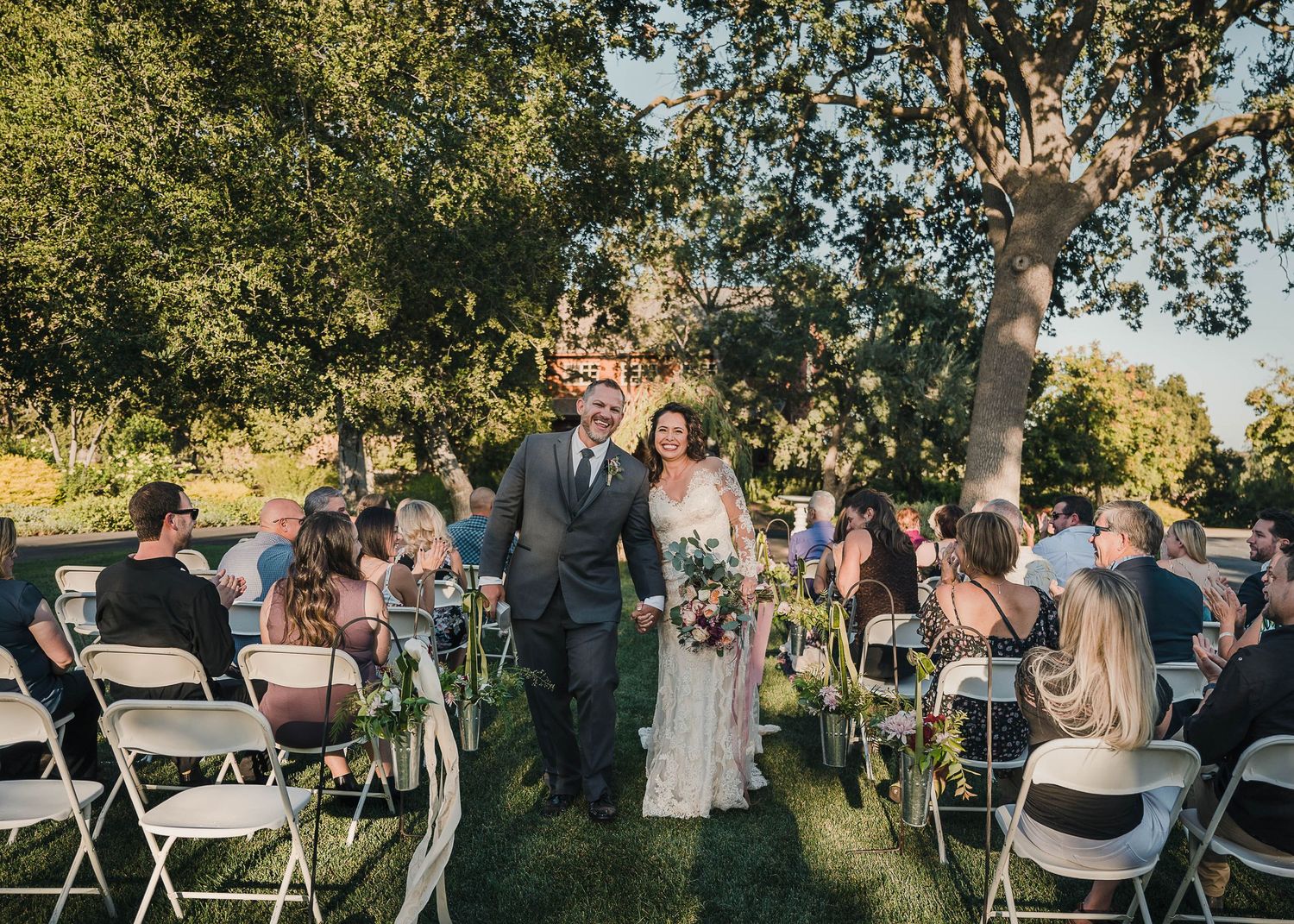 Newlyweds exit down an outdoor aisle lined with white chairs and excited wedding guests.