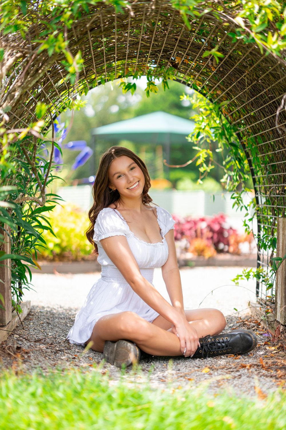 Person in white dress posing under garden archway surrounded by greenery and colorful flowers.