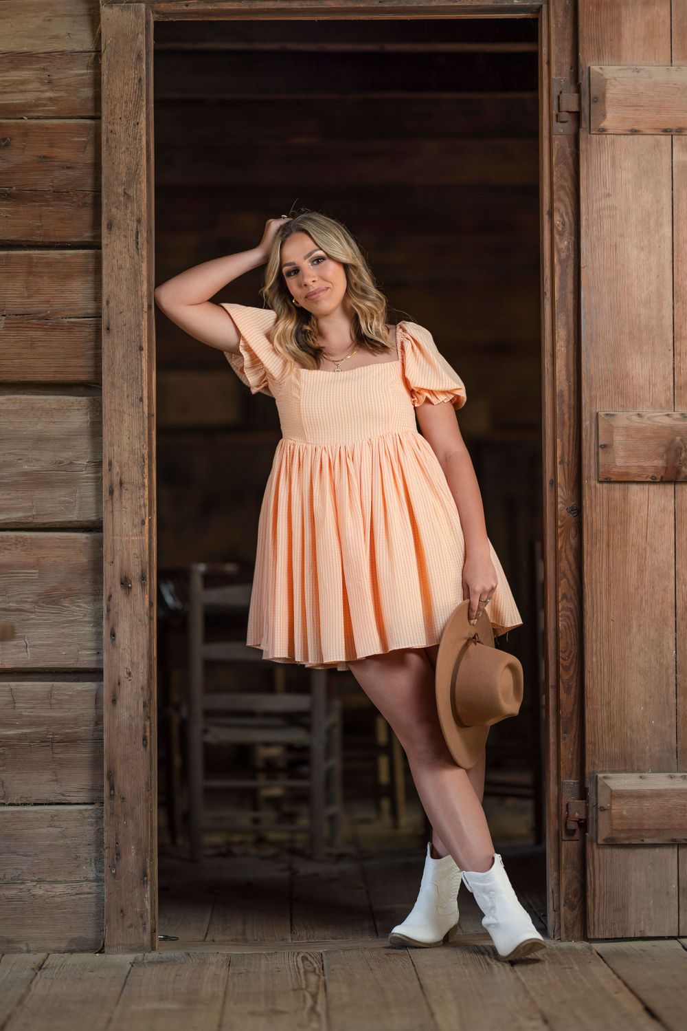 A person in a peach babydoll dress and white boots poses in a rustic wooden doorway.
