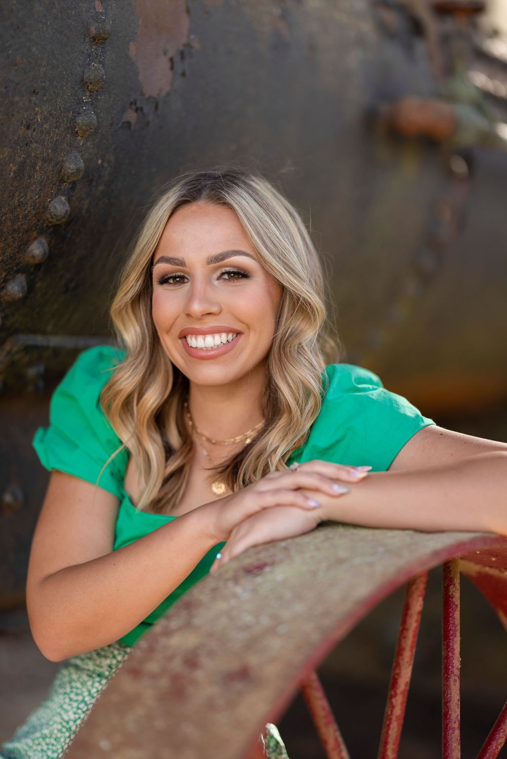A person in an emerald green top poses with wavy hair against a rustic metal background.