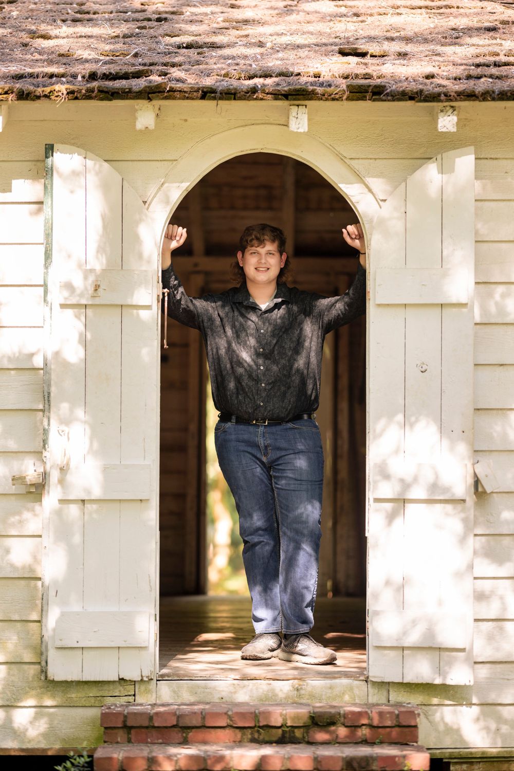 Person posing in doorway of white brick building wearing dark shirt and jeans.