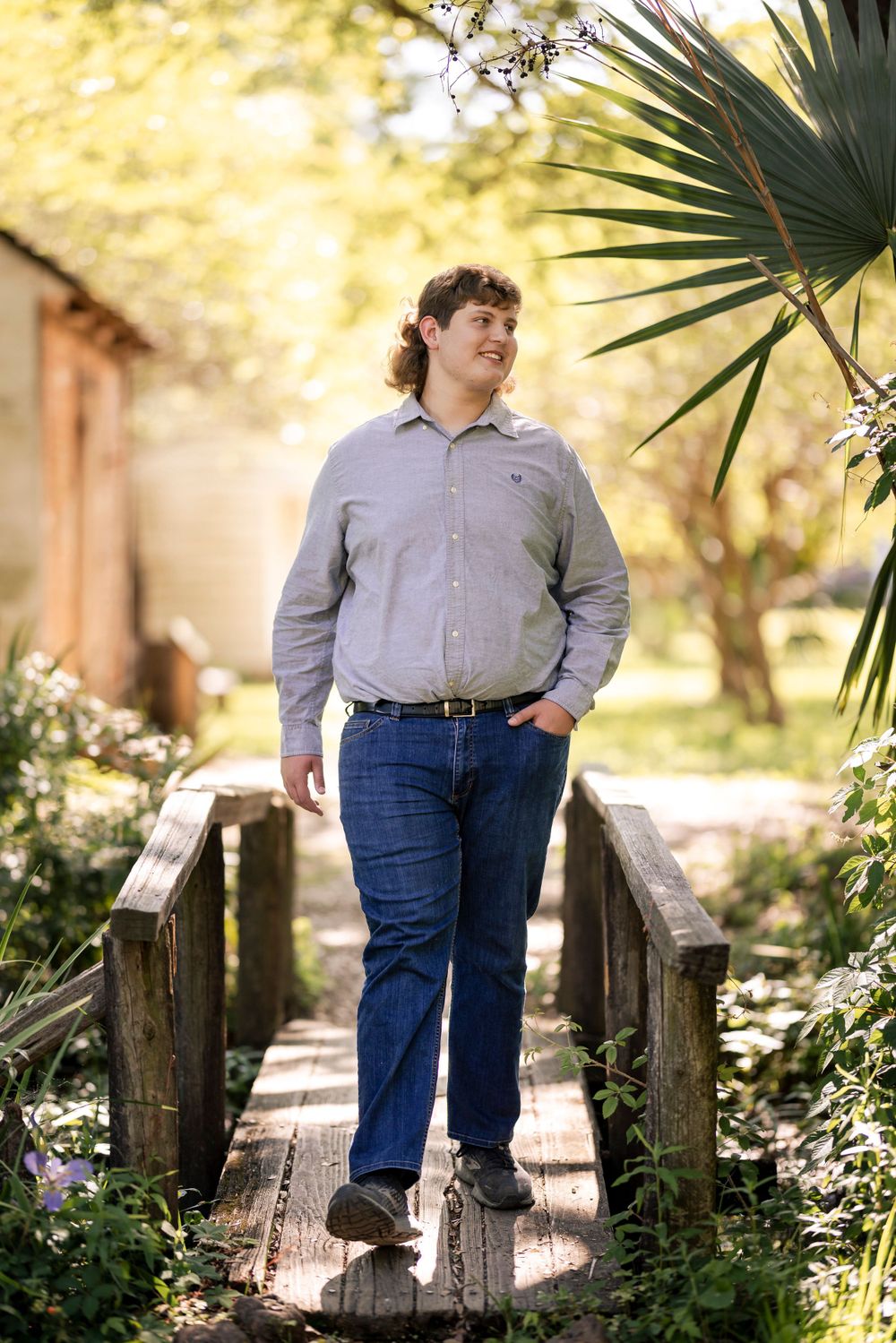 Person in grey shirt and jeans standing on wooden bridge in garden setting.