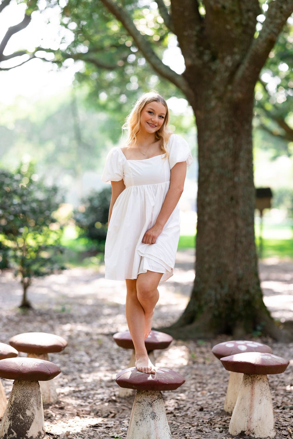 Person in white dress standing on decorative mushroom sculptures under large tree.
