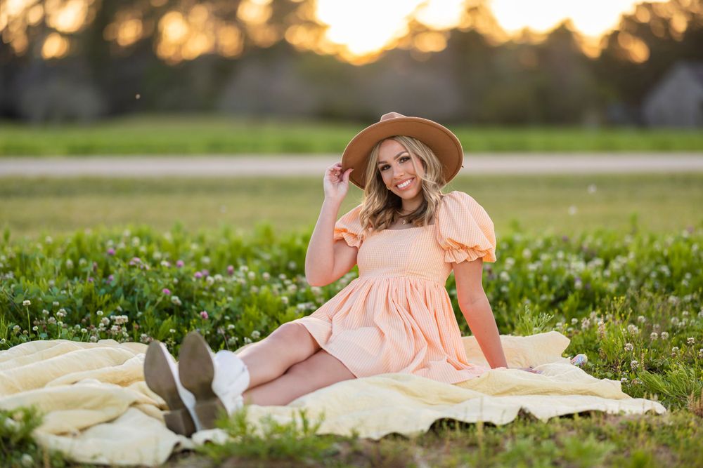 A person in a peach dress and sun hat poses on a blanket in a grassy field at sunset.