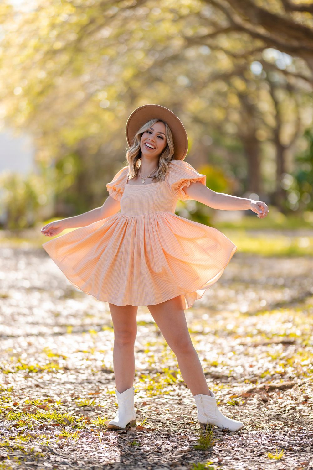 A person in a peach dress and brown hat twirls joyfully among fall leaves at sunset.