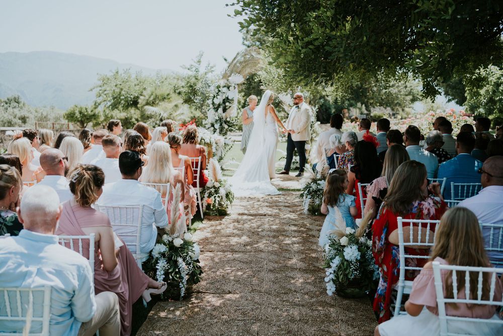 Outdoor wedding ceremony with white chairs lined up under trees and floral aisle decorations.