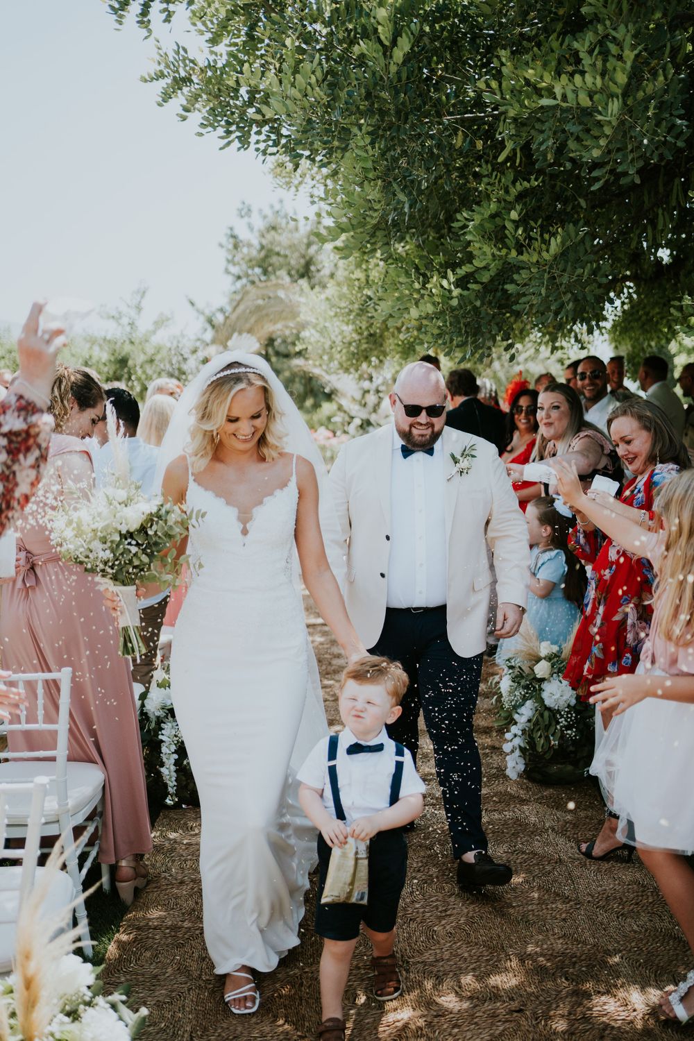 Wedding party walks down aisle with guests seated on white chairs under olive trees.
