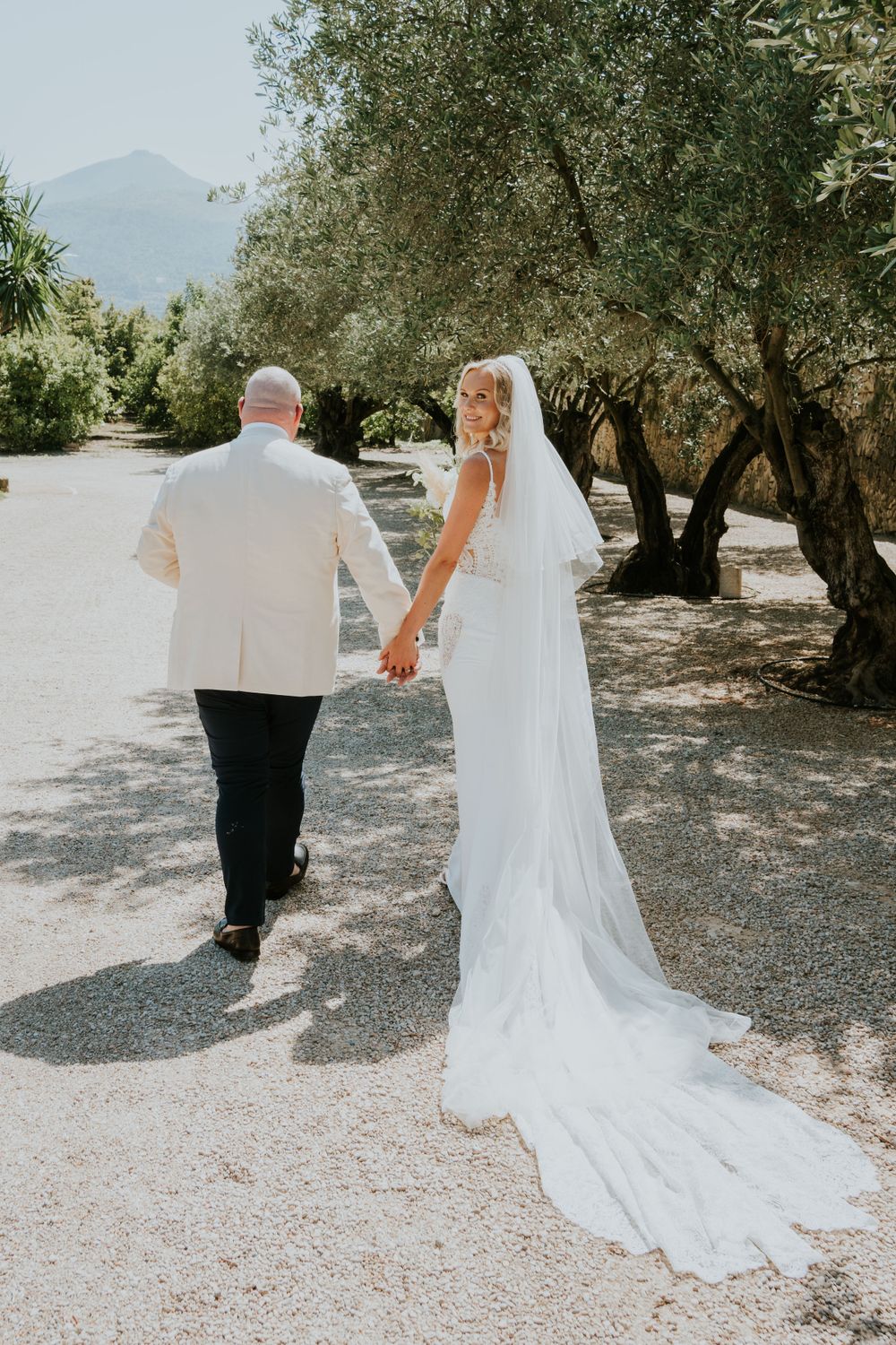 Couple walks hand in hand down sunlit path with bride's long veil trailing behind.