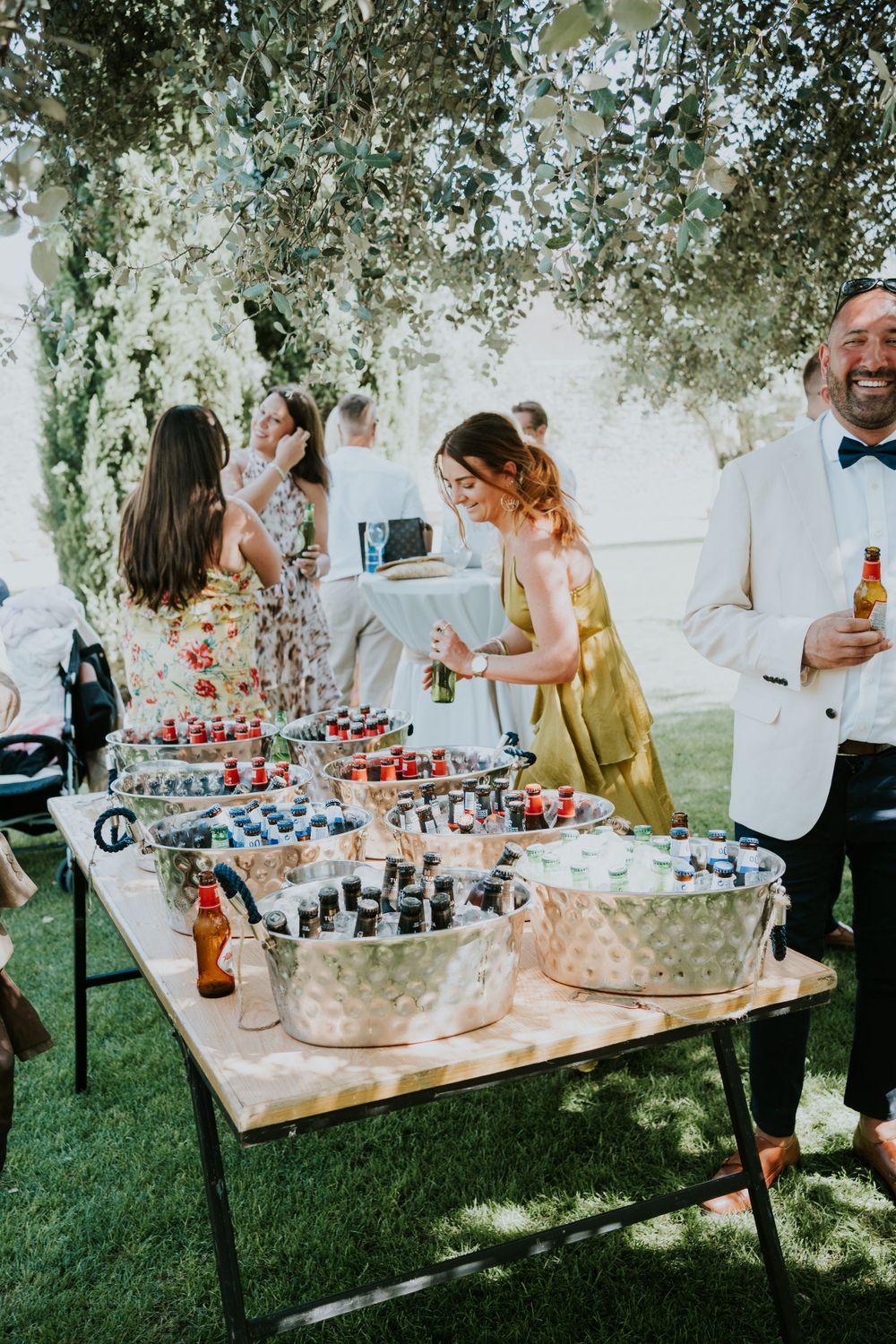 Wedding reception beverage station with drinks in ice buckets under shaded trees.