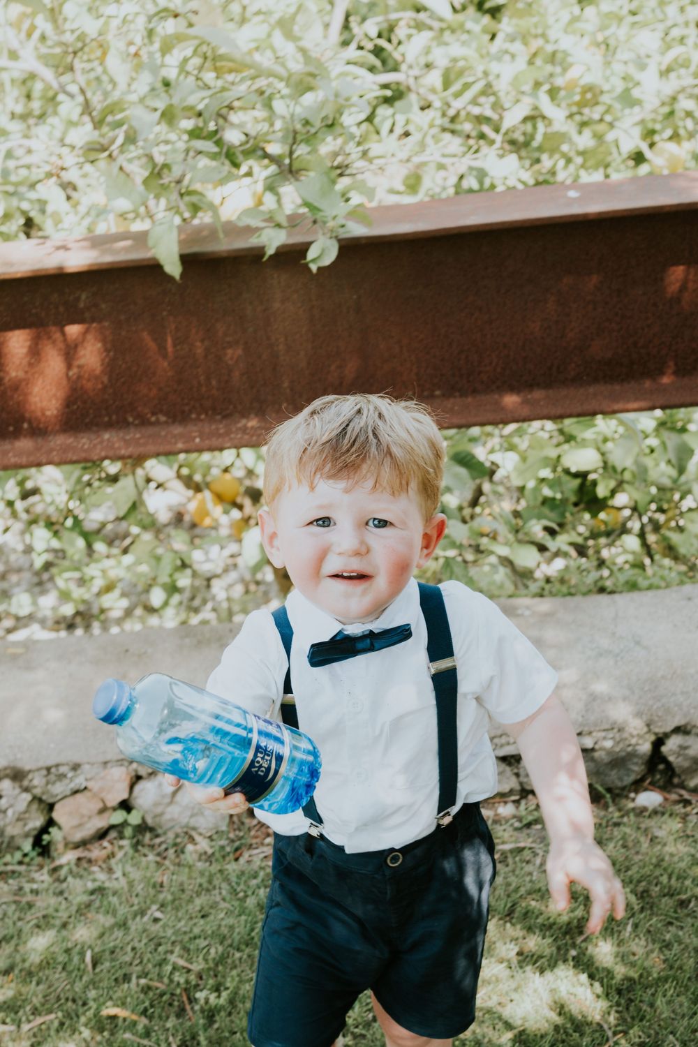 Young ring bearer in suspenders and bow tie carrying a blue water bottle at outdoor wedding.