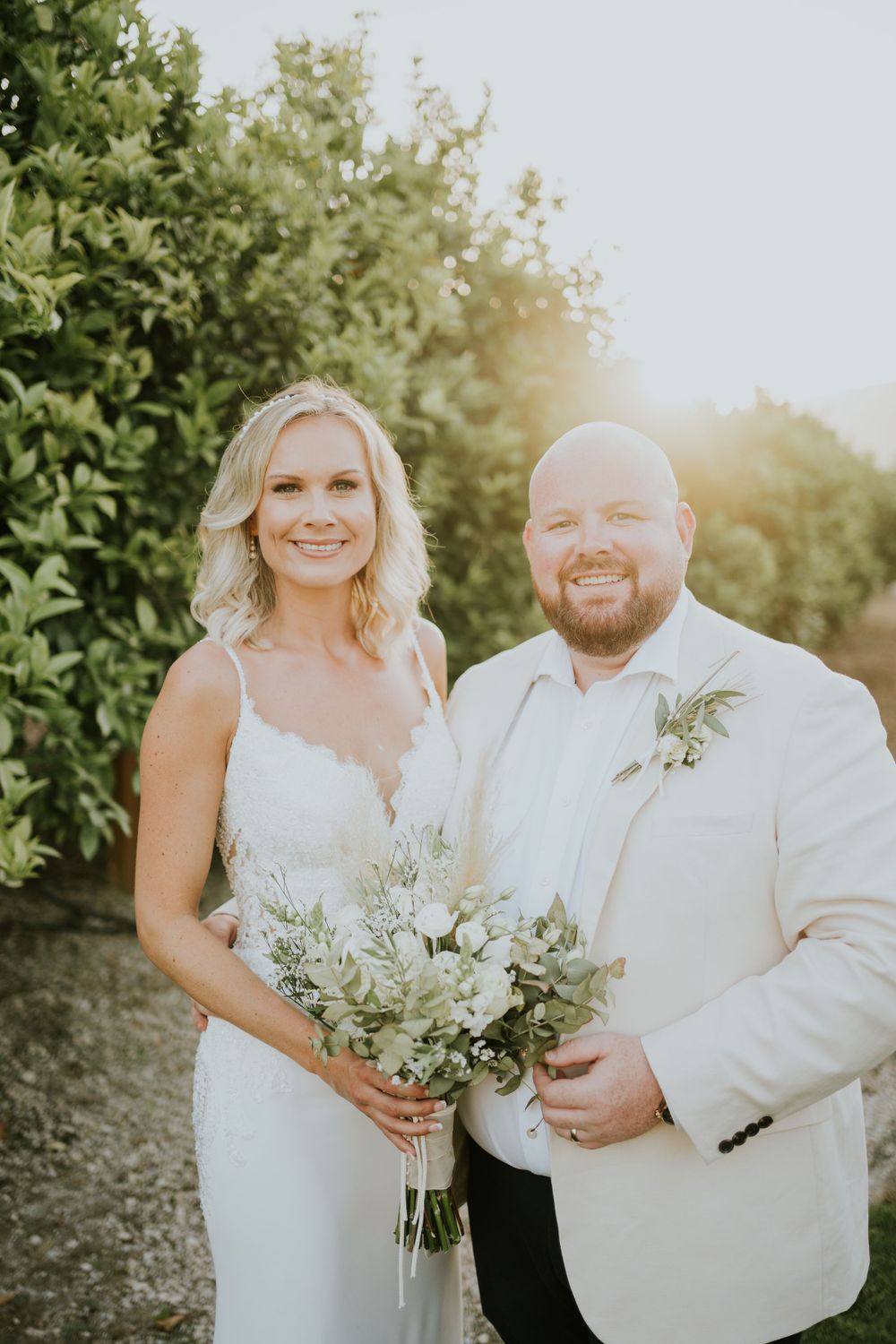 Newlyweds posing with bouquet against garden backdrop during sunset.