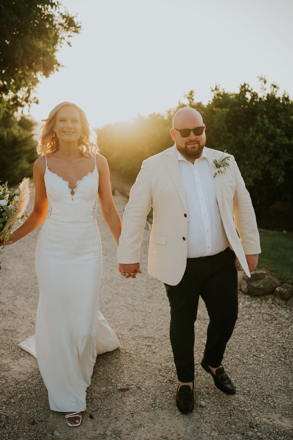 A couple walk hand in hand on a gravel path during sunset in their wedding attire.