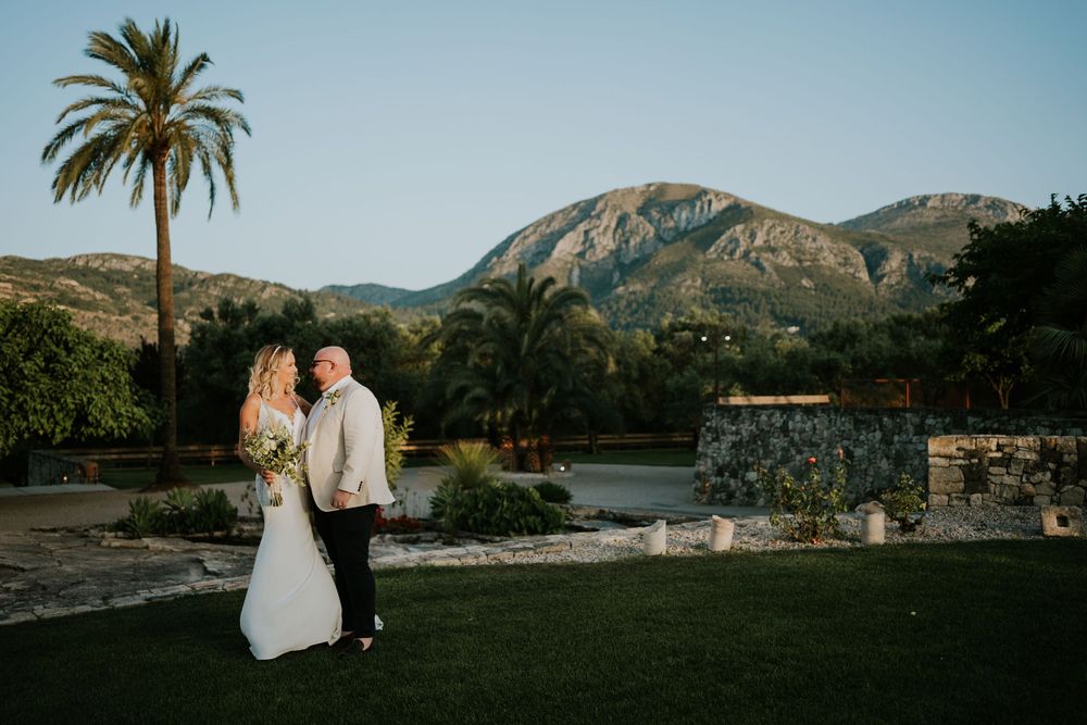 A couple in wedding attire pose against a scenic mountain landscape with a palm tree at sunset.