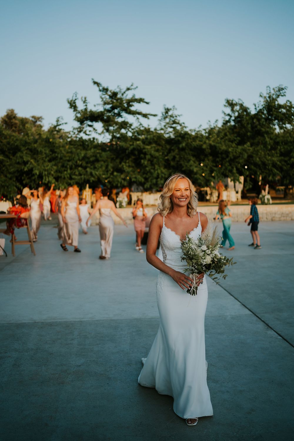 A bride in a fitted white dress holds a bouquet while standing on an outdoor patio at sunset.
