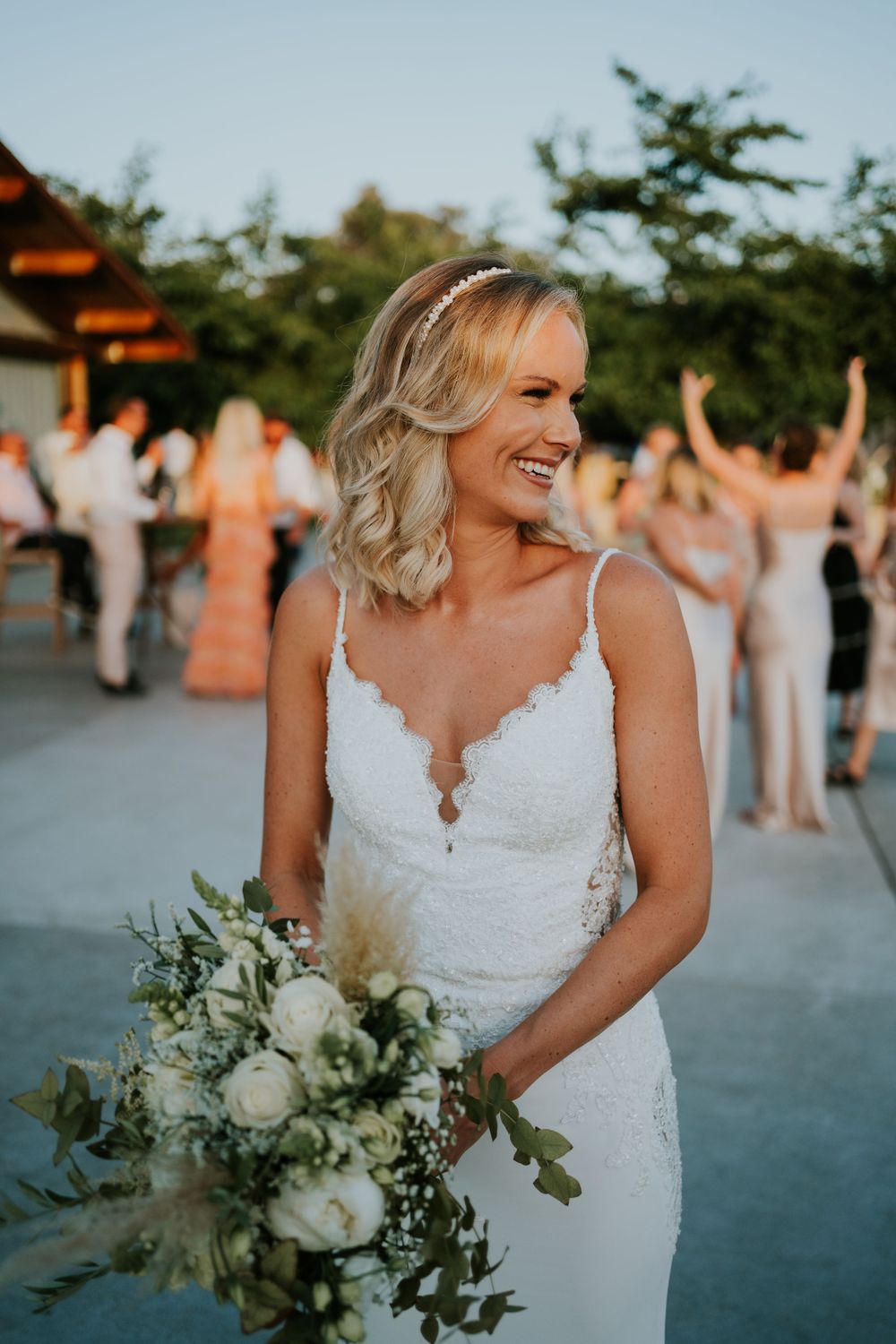 A bride holding a white floral bouquet smiles joyfully during an outdoor celebration.