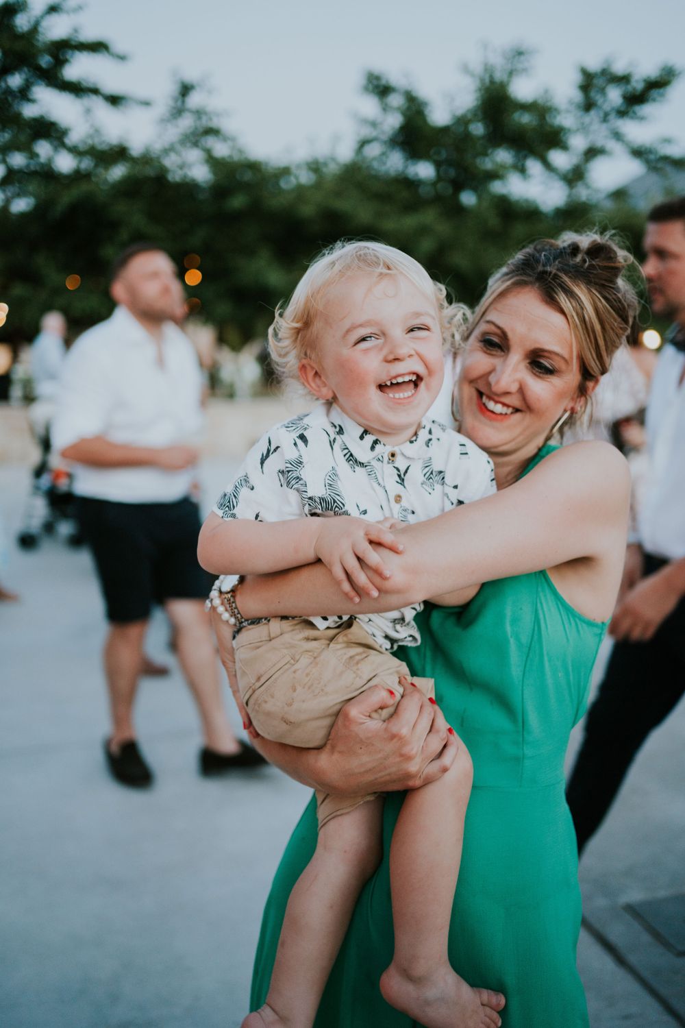 A guest in a green dress dances with a laughing child at an outdoor celebration.