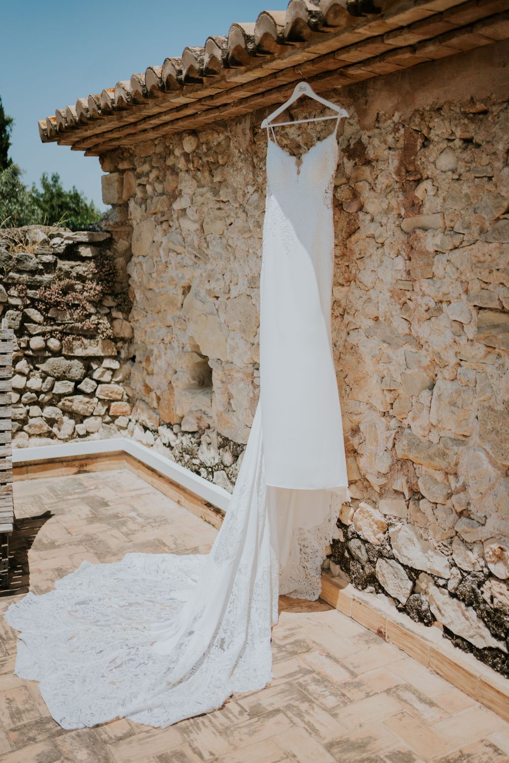 A white wedding gown with long train hangs against a rustic stone wall in Mediterranean setting.