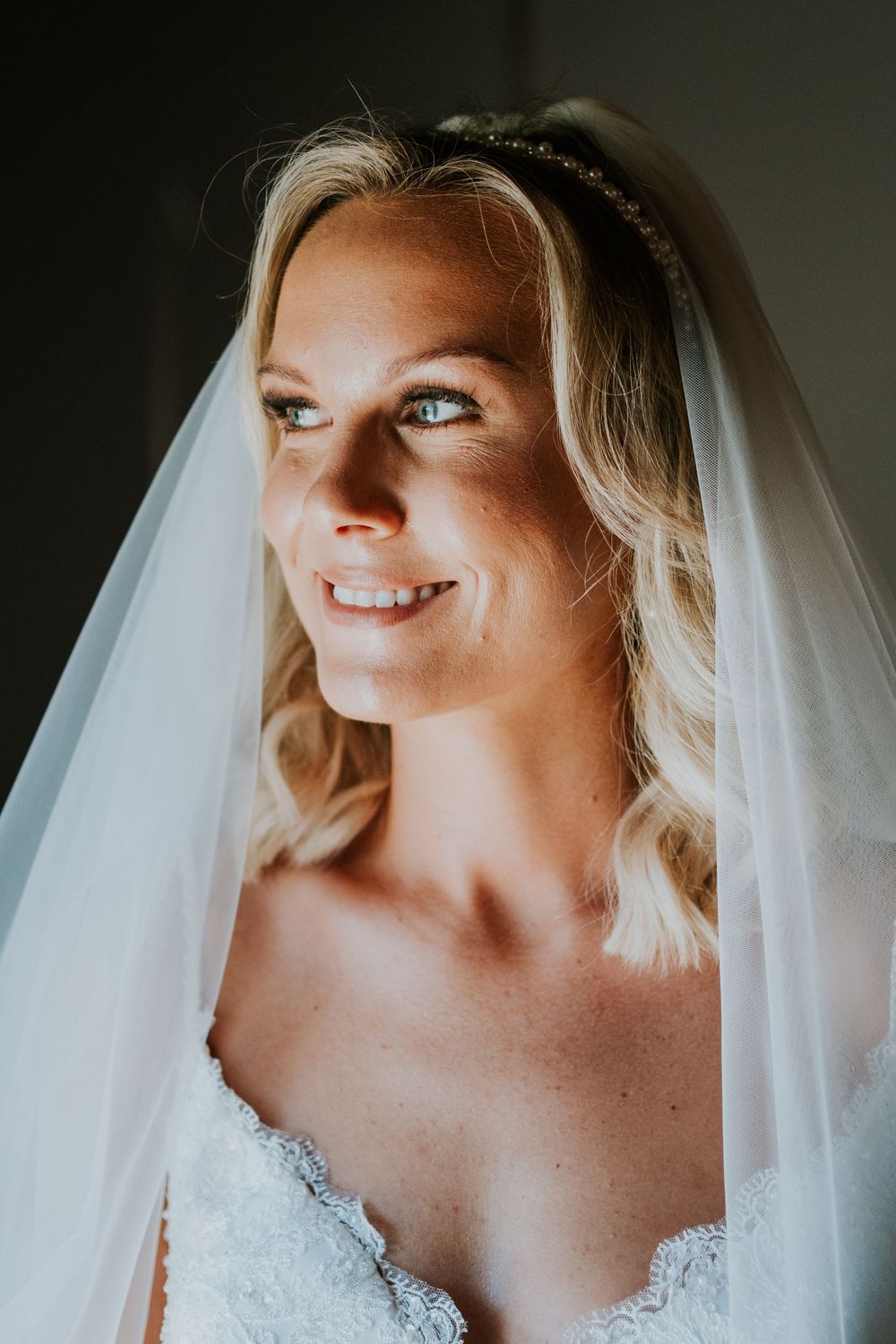 A bride in a white lace dress and veil smiles in a moody portrait on her wedding day.