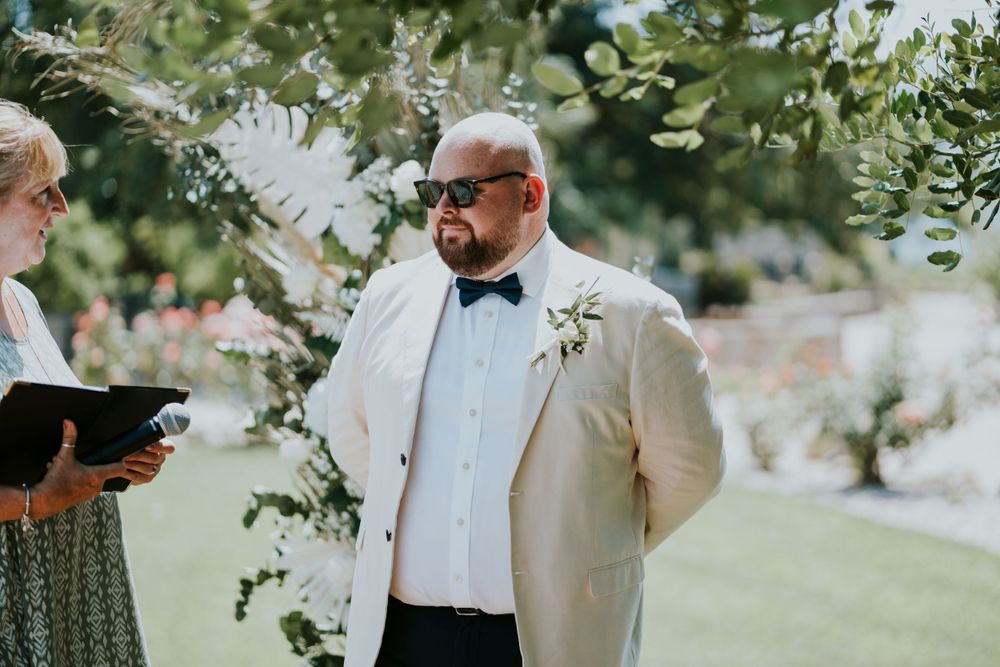Outdoor wedding ceremony with officiant and groom in white suit under tree branches.