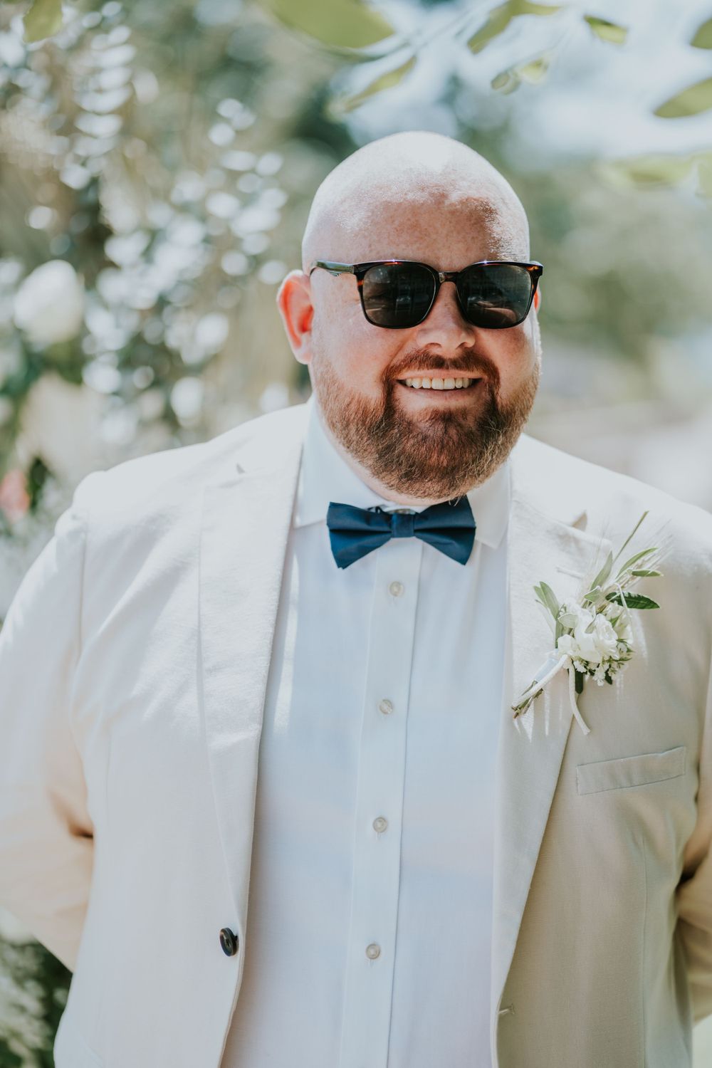 Groom in white suit jacket and blue bow tie smiles while wearing sunglasses outdoors.