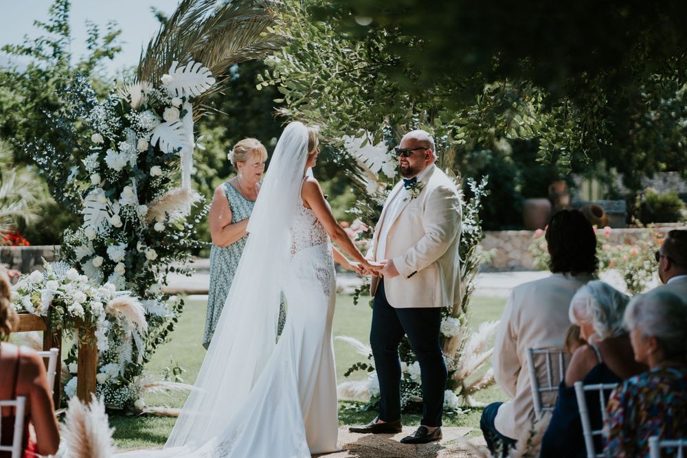 Outdoor garden wedding ceremony with floral arch and guests seated on white chairs.