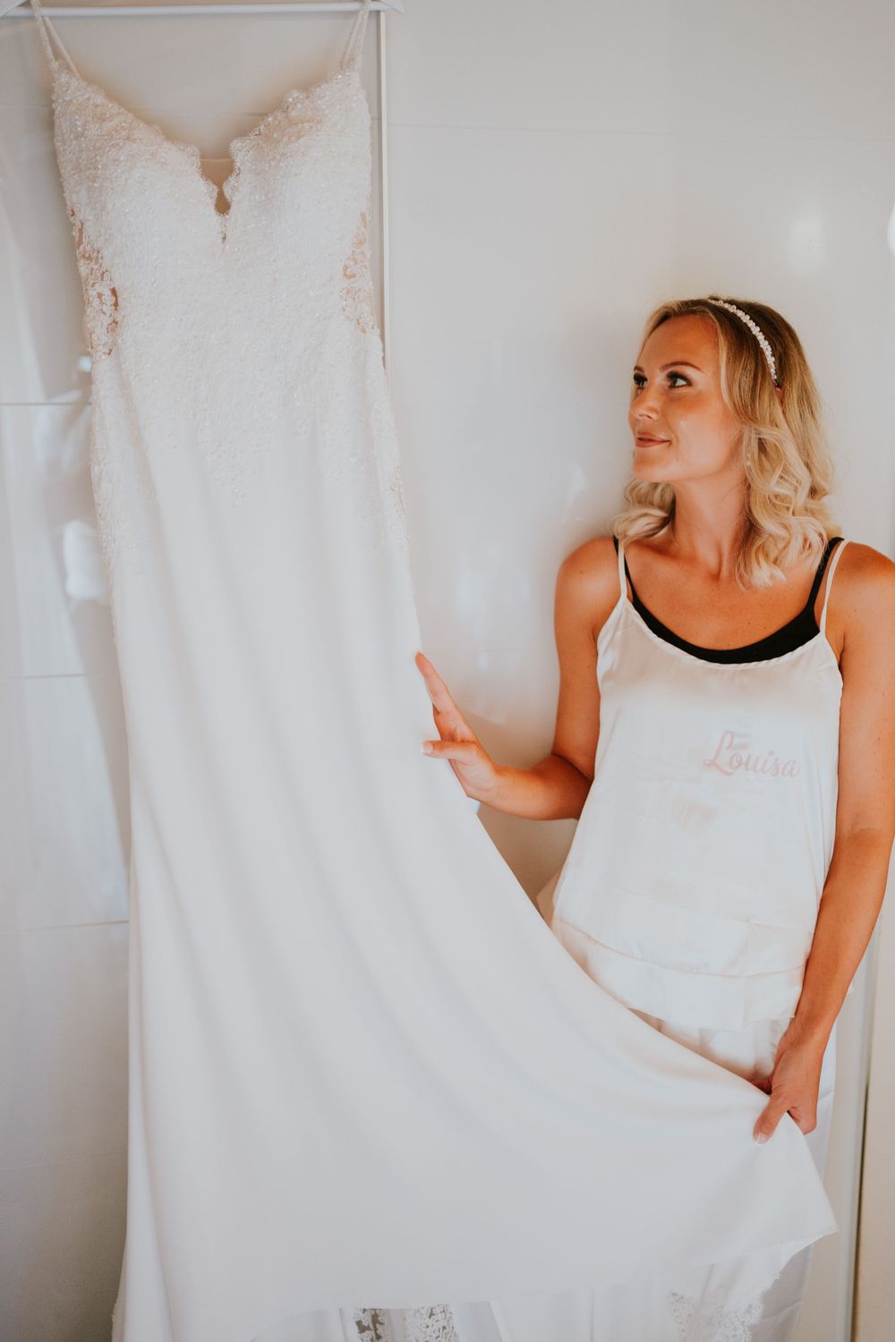 A elegant white lace wedding dress hangs on display as someone in a white tank top admires it.