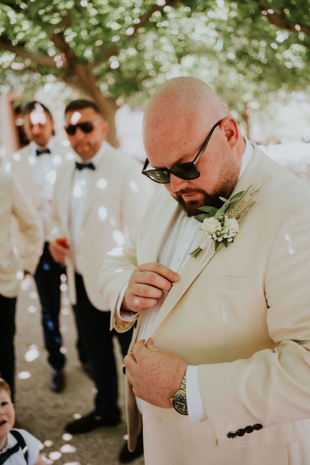 Wedding party member in cream suit adjusts boutonniere during outdoor celebration.