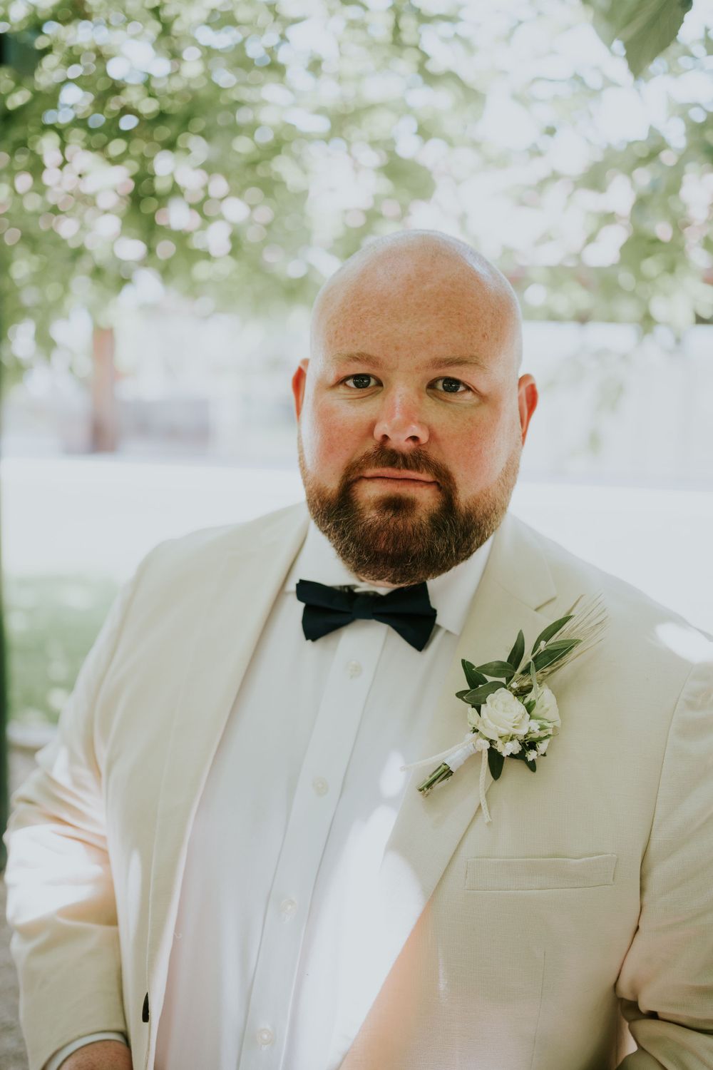 Groom in white tuxedo with black bow tie and floral boutonniere poses outdoors.