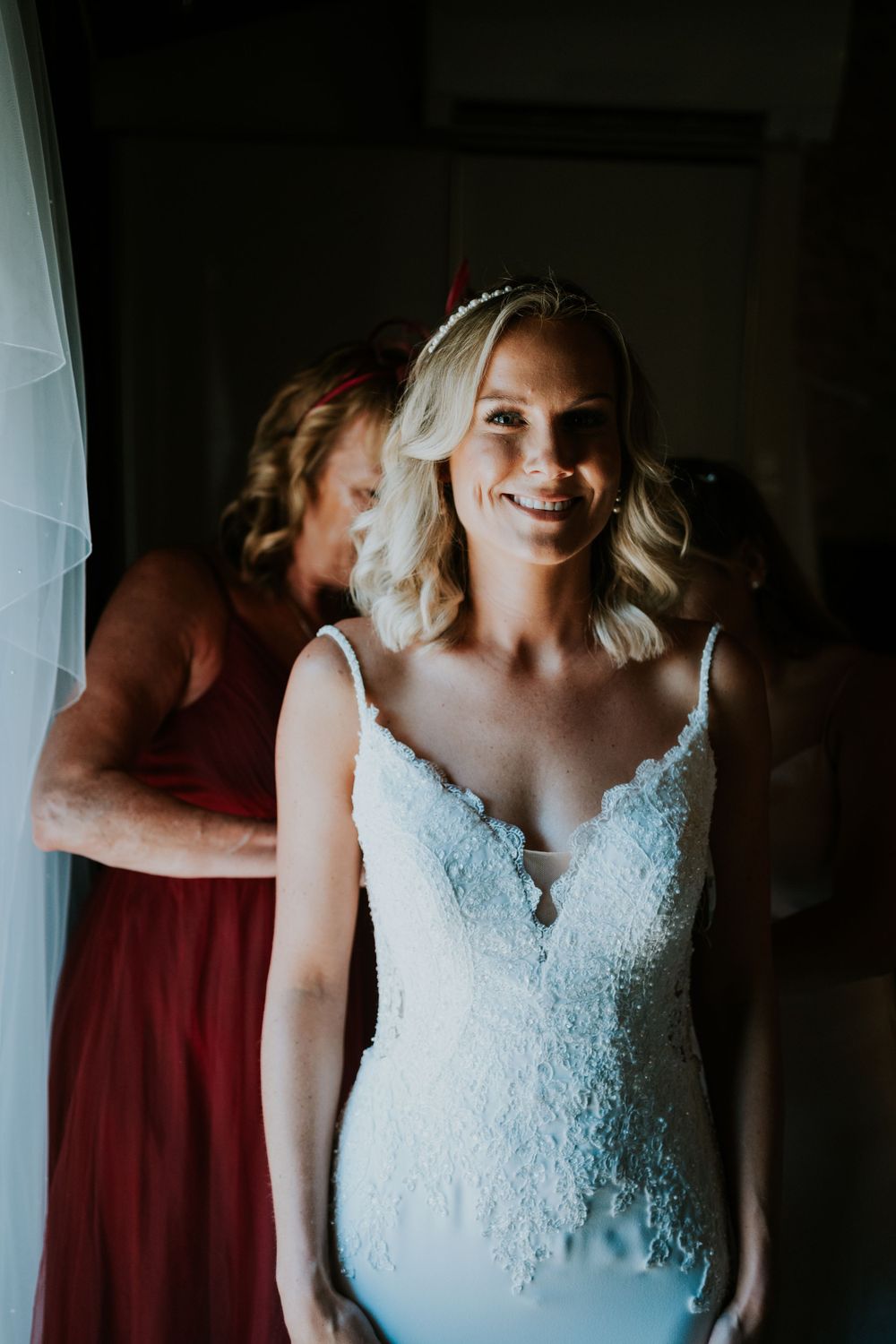 Wedding dress fitting moment captured in dramatic lighting with bridesmaids in coral dresses.