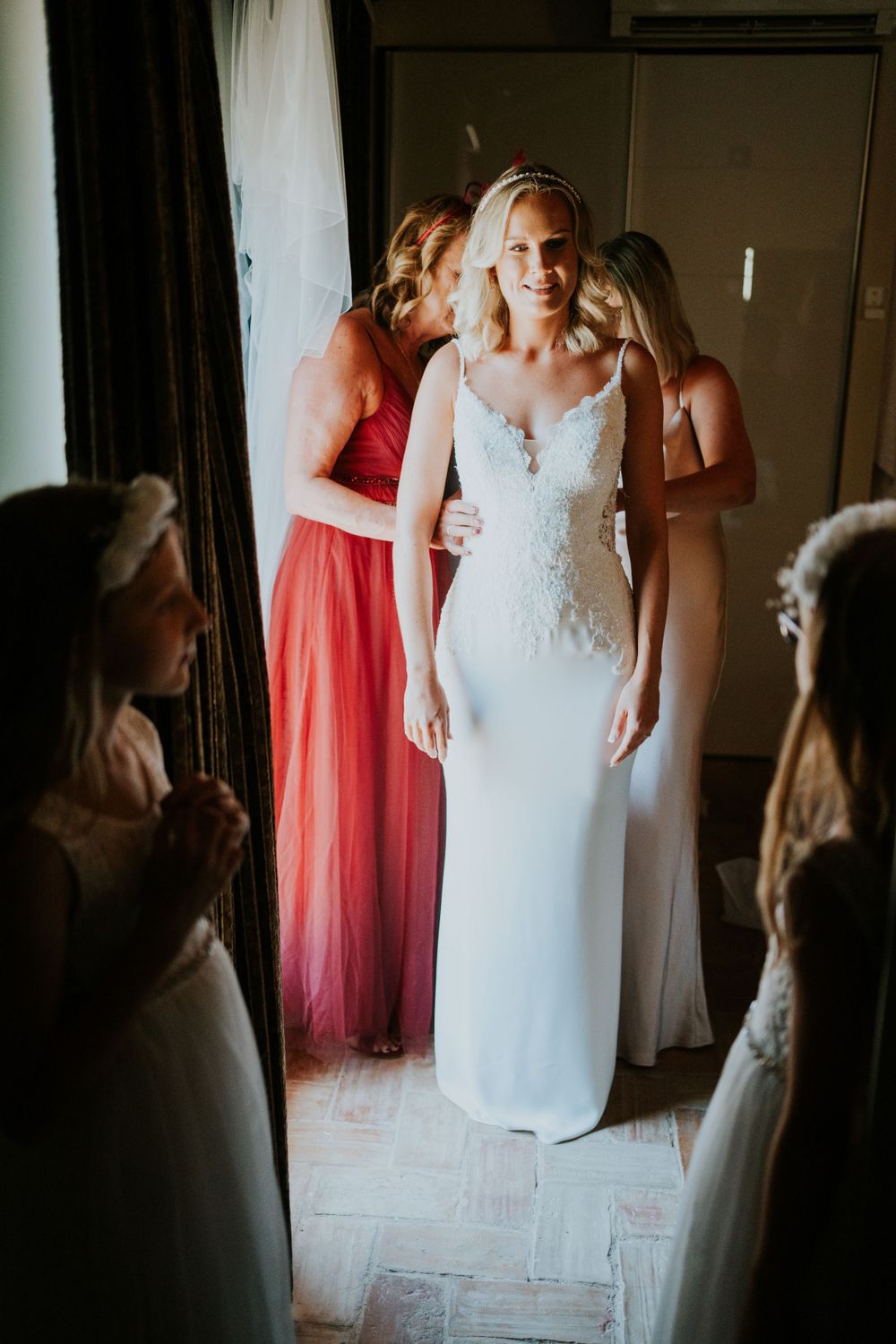 Bride in fitted lace gown surrounded by bridesmaids in coral dresses during pre-wedding preparations.