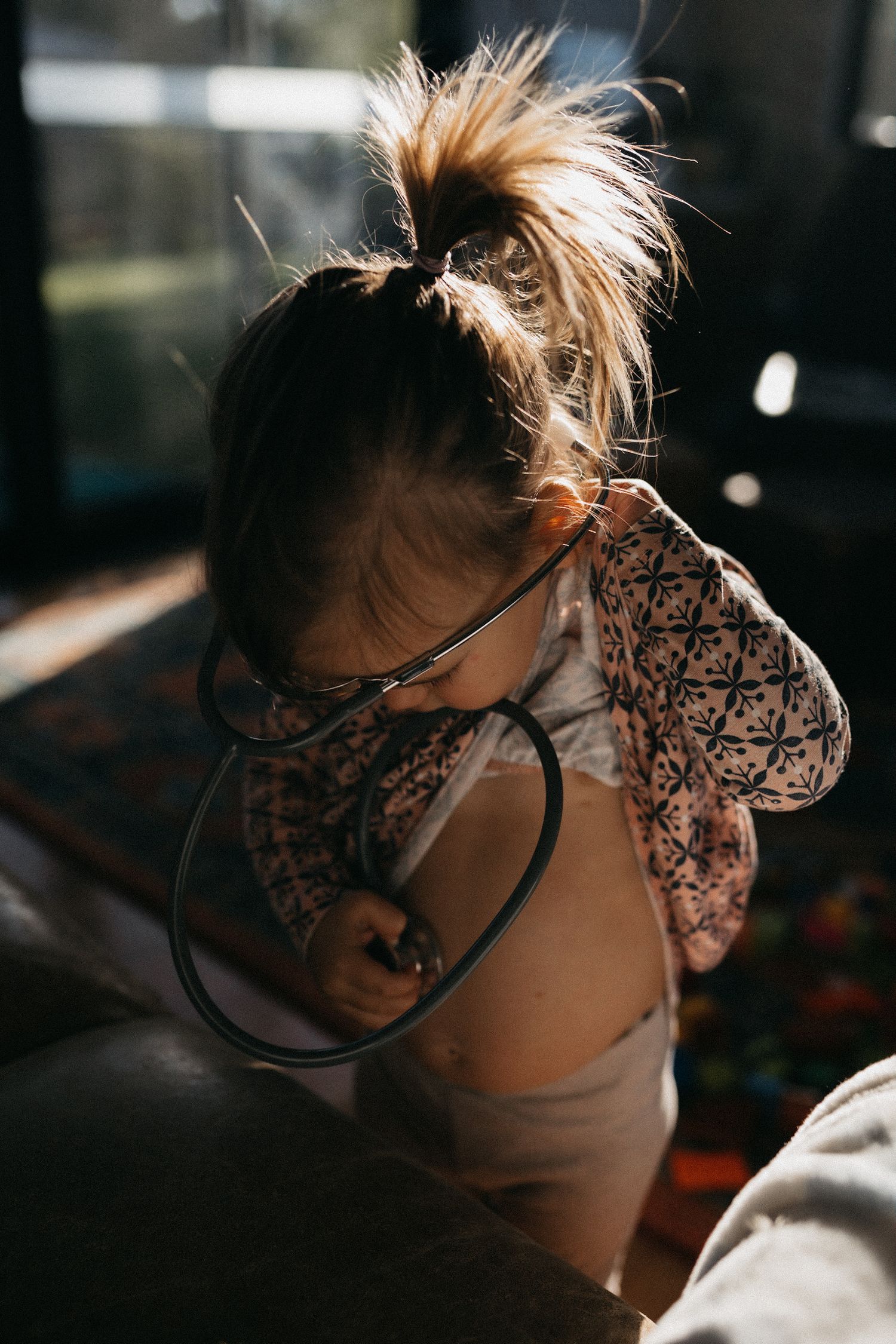 A toddler playing doctors, with a stethoscope against her tummy, at home