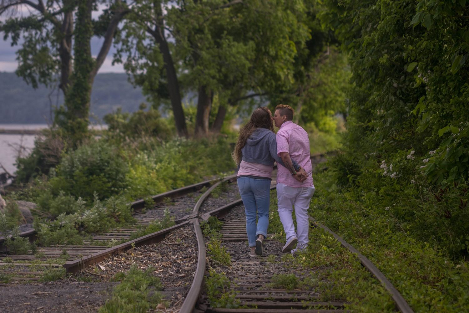 Couple walks together on abandoned railroad tracks surrounded by green foliage.