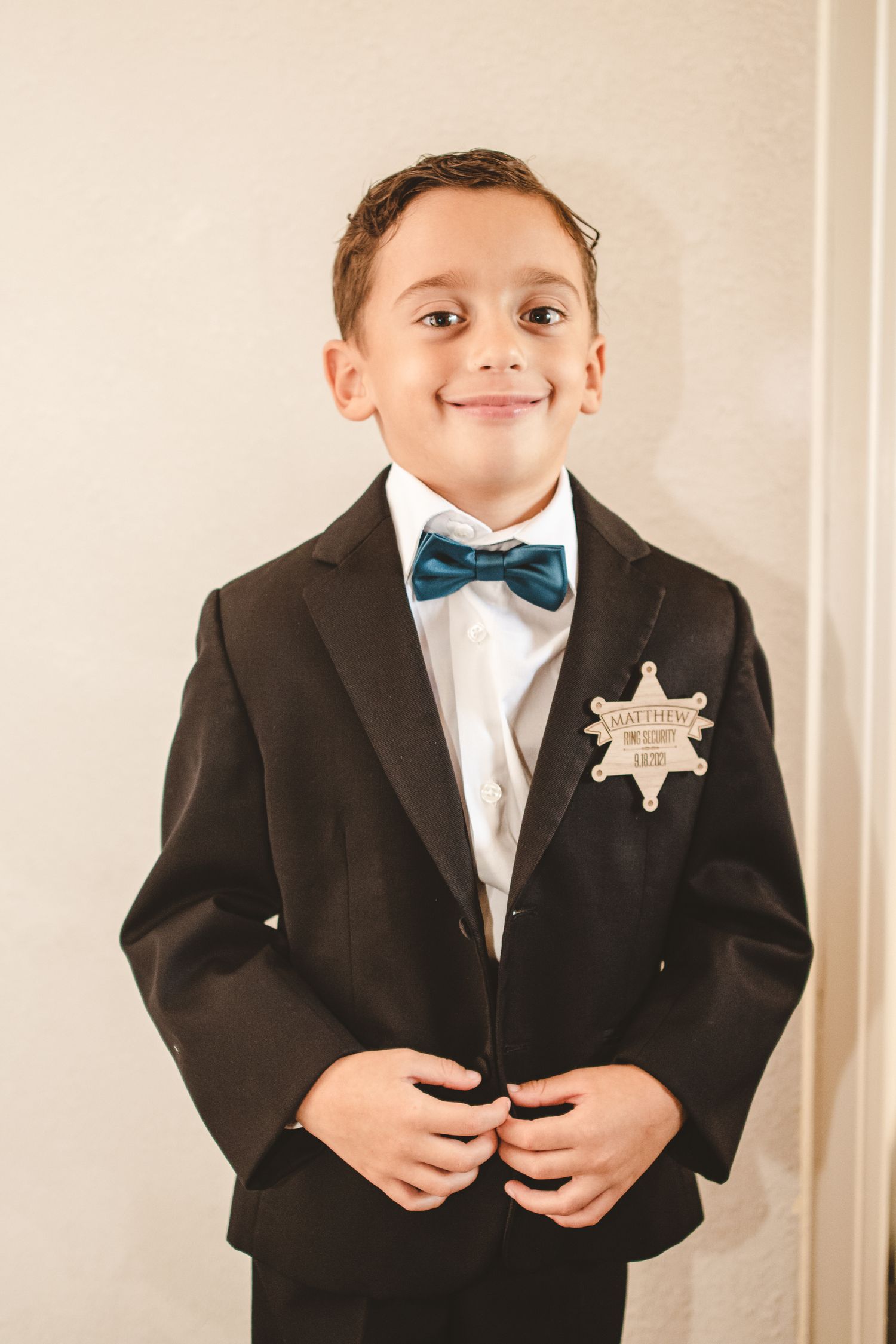 Young child in formal black suit and blue bowtie posing against beige background.