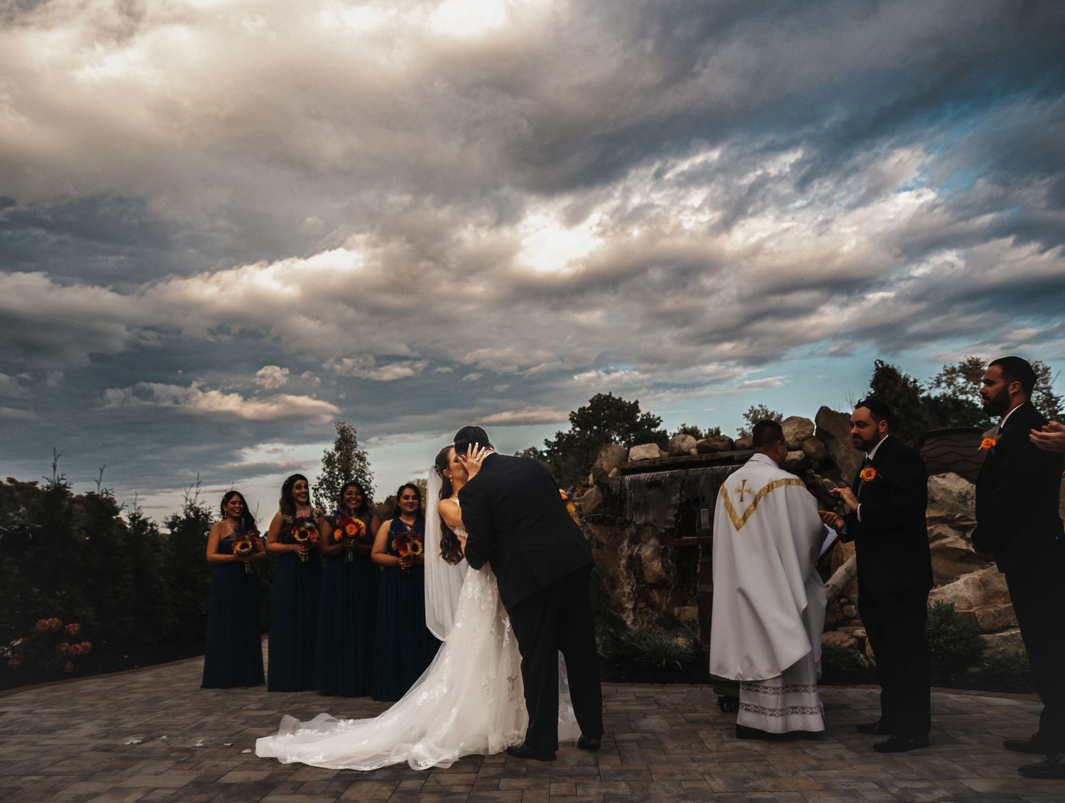 Wedding ceremony taking place outdoors under dramatic cloudy sky at sunset.